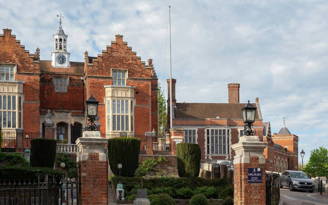 The Druries gateway at Harrow School on a bright day, with red-brick gate pillars topped by black lanterns and iron railings in front of the stepped-gable brick building and its white clock tower