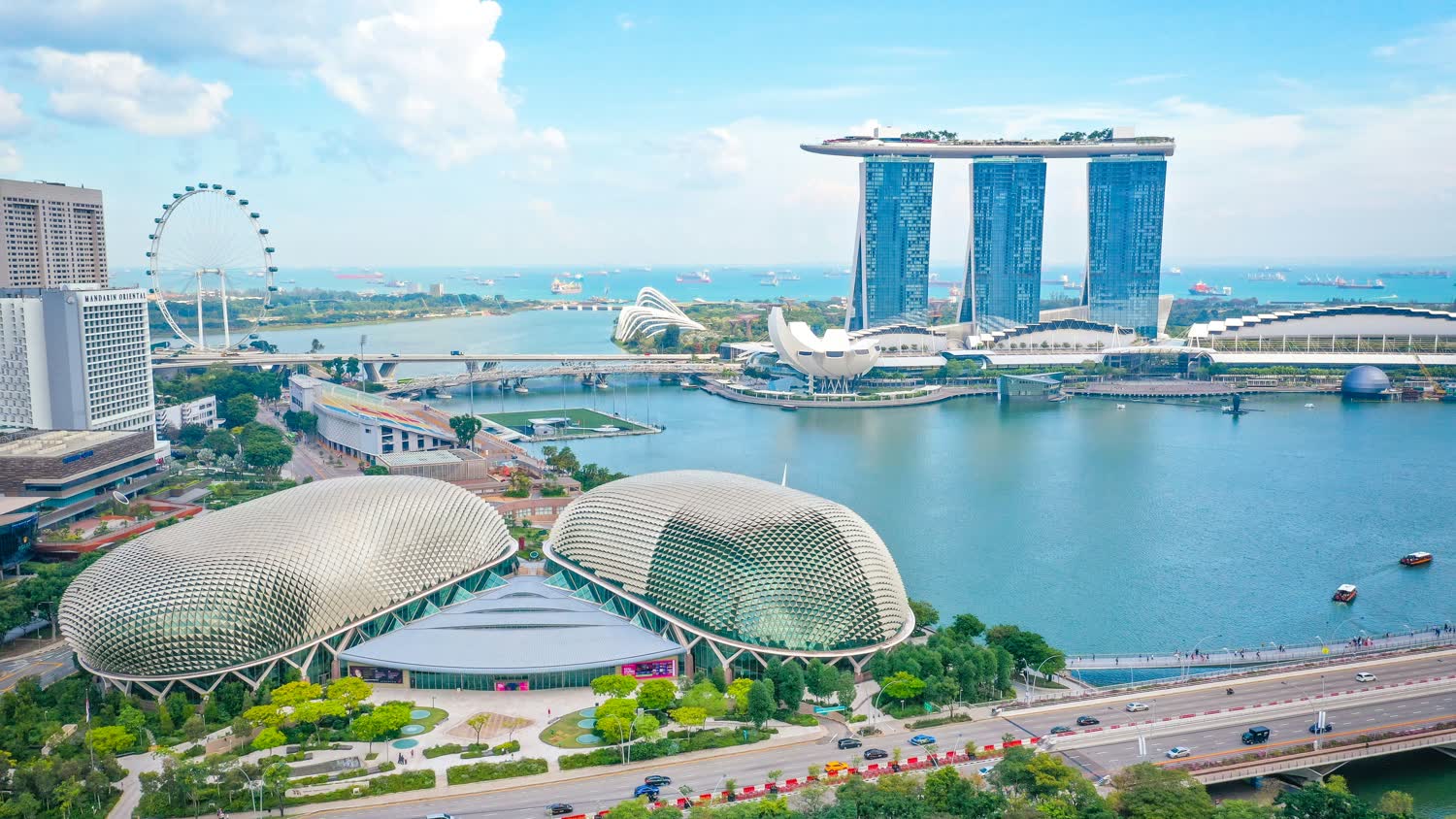 Singapore seen from above on a clear day, with the twin domed roofs of the theatre on the bay in the foreground, the observation wheel and the waterfront hotel beyond, and ships on the horizon
